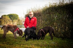 Una persona con una giacca rossa è inginocchiata sull'erba, circondata da quattro cani che annusano il terreno vicino a una siepe.