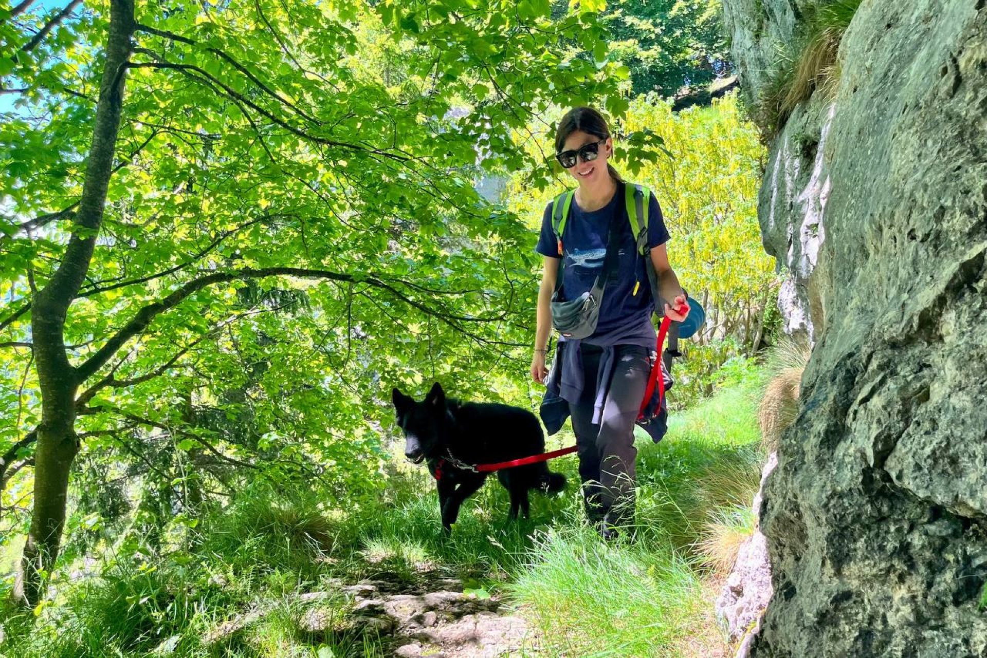 Una donna che indossa occhiali da sole e uno zaino porta a spasso un cane nero al guinzaglio lungo un sentiero boscoso accanto a una scogliera rocciosa.